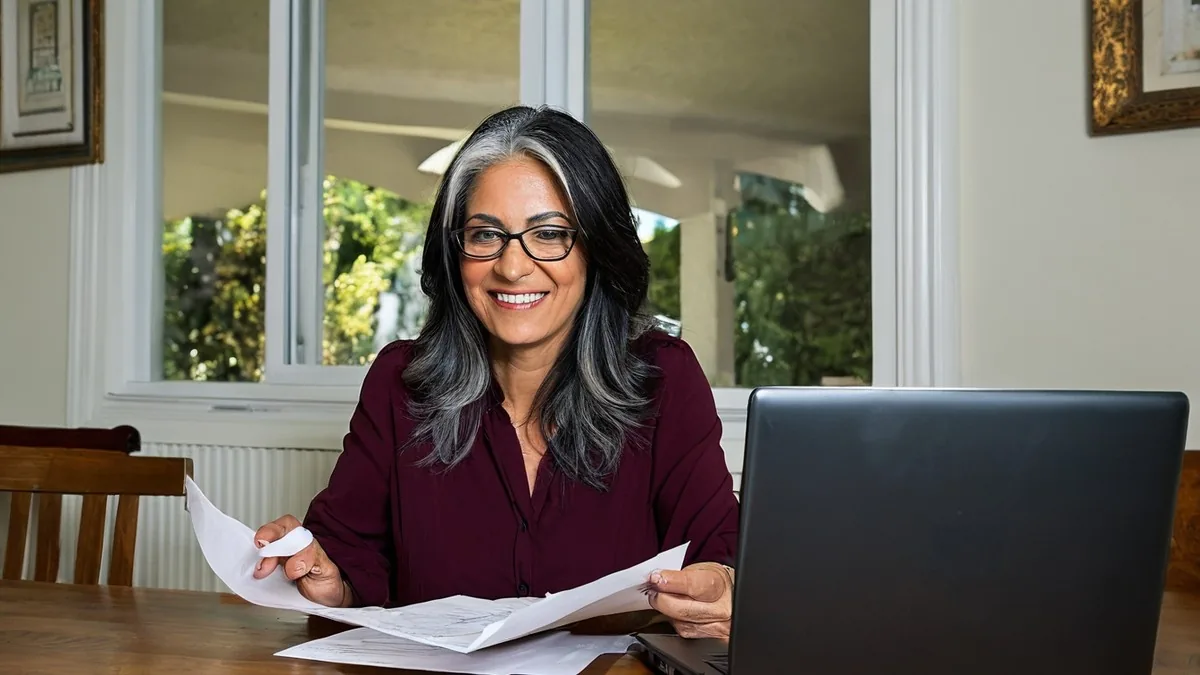 Smiling woman with long silver-streaked dark hair and glasses reviewing paperwork beside a laptop